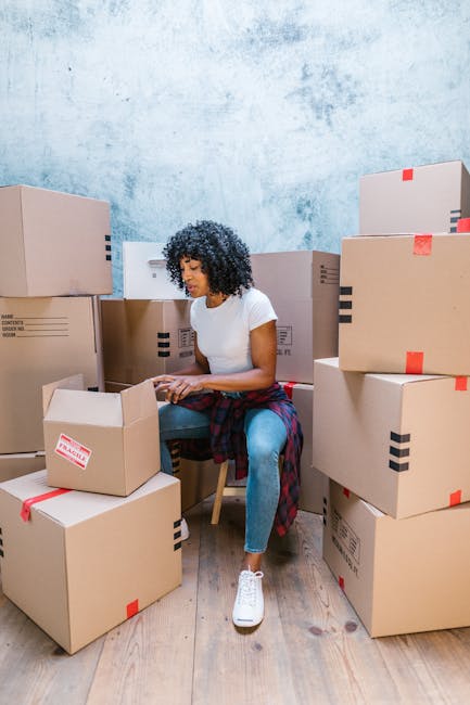 A man with an afro hairstyle and a beard wearing a blue T-shirt is standing inside a property, surrounded by moving boxes and packaging materials. He is holding a clipboard and writing notes, likely a